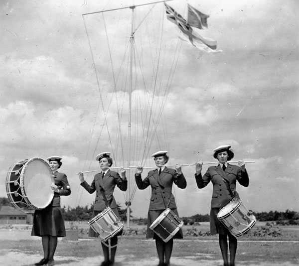 Photographie en noir et blanc – Quatre femmes en uniforme de la marine sont placées en rang. Celle à gauche joue de la grosse caisse. Les autres, à leur caisse claire, tiennent leurs baguettes à l’horizontale. On aperçoit derrière le Red Ensign.
